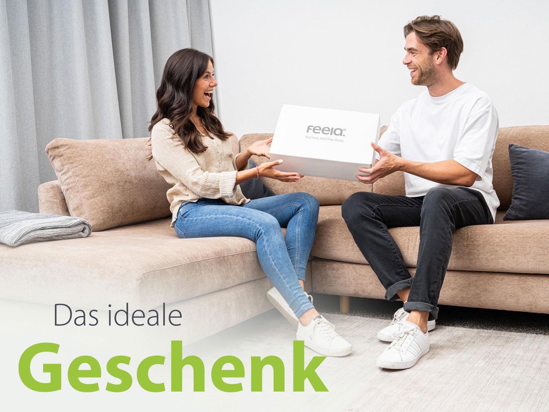 Bild eines lächelnden Paares auf einem Sofa in einem modern eingerichteten Wohnzimmer. Der Mann übergibt der Frau eine weiße Geschenkbox mit dem feela® Logo. Text unten auf dem Bild: 'Das ideale Geschenk'.