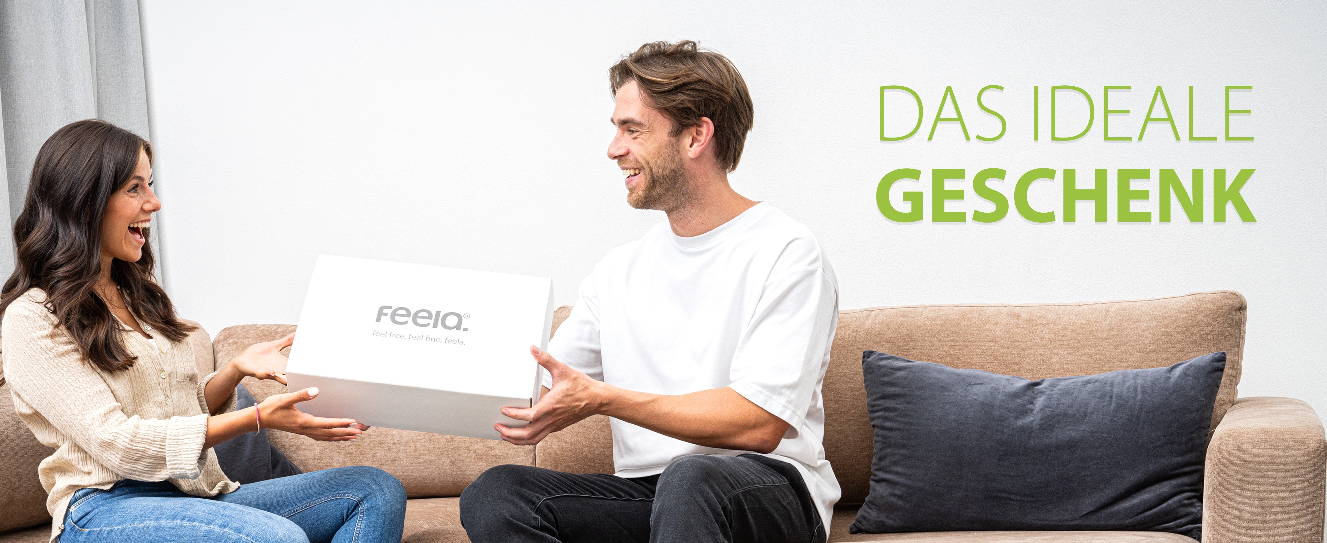 Eine Frau überreicht einem Mann eine Geschenkbox von feela, beide lächeln und sitzen auf einem Sofa, unterstrichen von dem Text 'Das ideale Geschenk', was auf das orthopädische Auto-Sitzkissen von feela als eine ausgezeichnete Geschenkoption hinweist.