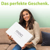 Strahlende Frau hält eine elegante weiße Geschenkbox von feela, mit dem Slogan 'Das perfekte Geschenk.' auf einem grünen Hintergrund, ideal zum Verschenken von Wohlbefinden und Komfort.