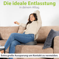 Frau entspannt auf dem Sofa mit erhobenen Armen, sitzend auf einem ergonomischen Kissen von feela, das durch seine extra große Aussparung Druckkontakt vermeidet.