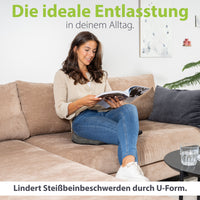 Entspannte Frau sitzt auf einem Sofa und liest ein Magazin, wobei sie ein U-förmiges Kissen benutzt, das als ideale tägliche Entlastung für Beinbeschwerden dargestellt wird.