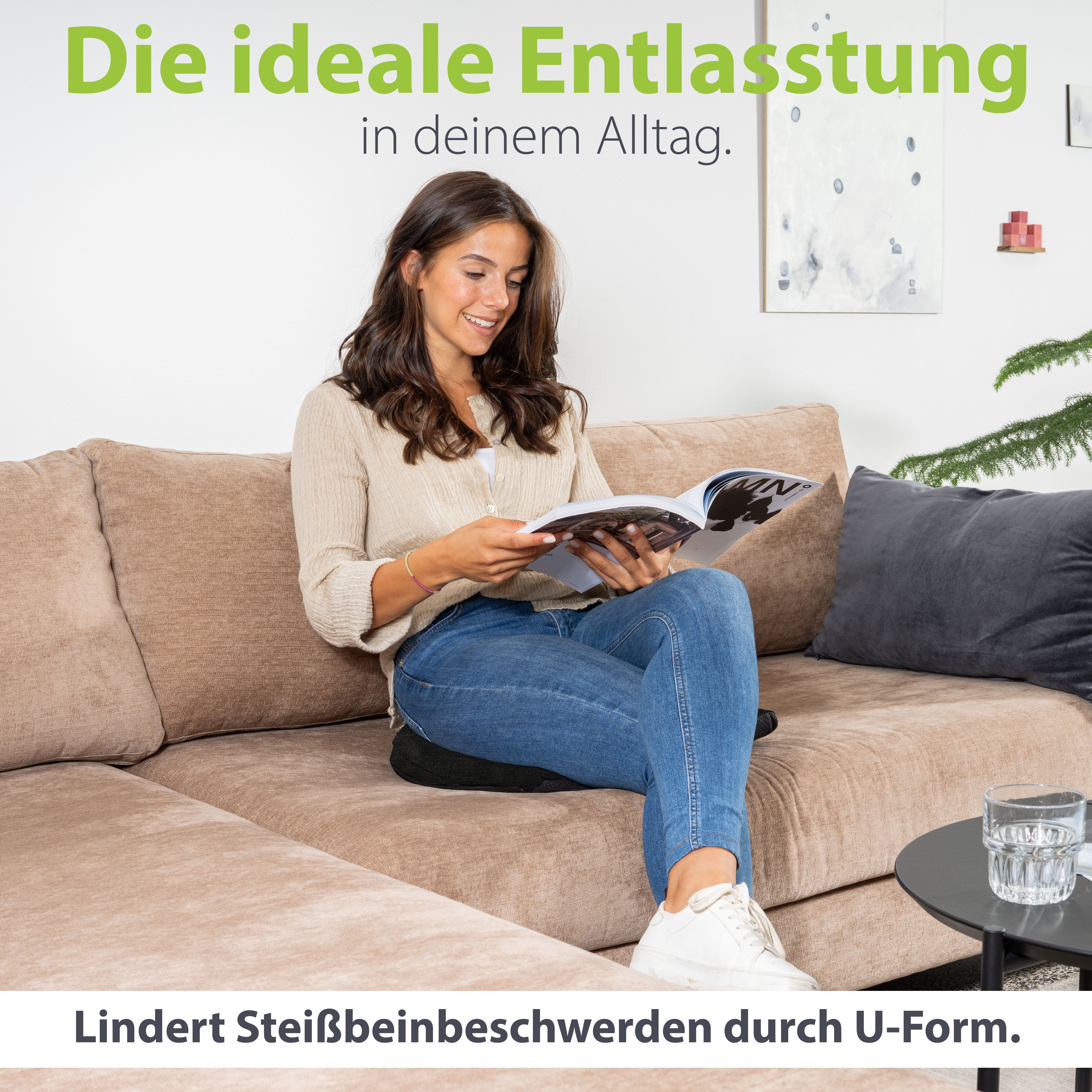 Entspannte Frau sitzt auf einem Sofa und liest ein Magazin, wobei sie ein U-förmiges Kissen benutzt, das als ideale tägliche Entlastung für Beinbeschwerden dargestellt wird.